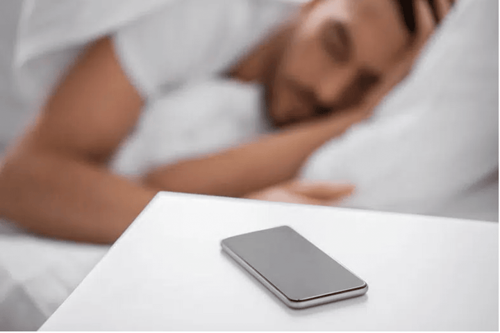 man laying down in bed with his head on his pillow, this part of the image is blurred because it is in the background while the cell phone on his nightstand appears in focus as the main subject