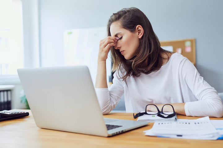 woman sitting at a desk with laptop, papers, and glasses on desk. She is pinching her head right below her eyes and near her nose indicating she has a headache. It is daytime, she is in an office.