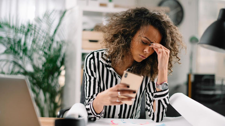 image of woman with a cell phone in her hand, in an office setting holding her head like she has a headache