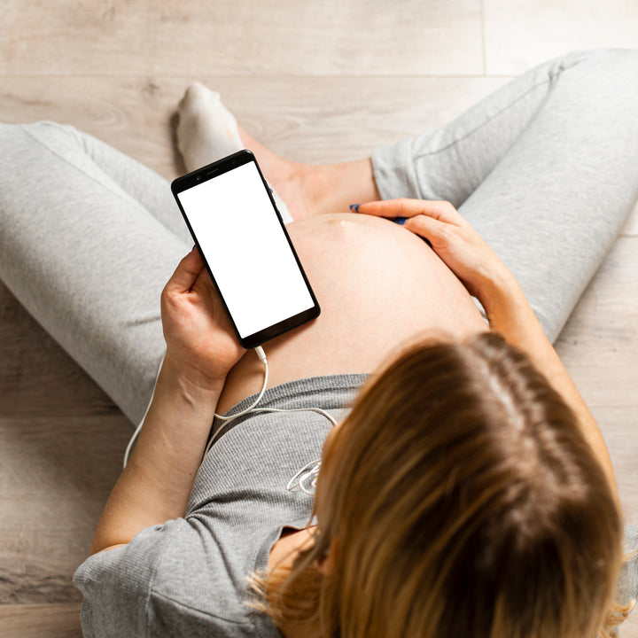 top view of a pregnant woman holding a cell phone in her left hand right up against her pregnant belly. She is sitting criss cross on the floor. The cell phone has a cable coming out of the bottom of it. 